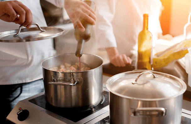 Closeup hands of unrecognizable chef pouring wine into pot with meat. In the background is a blurred figure of another chef.  - 写真・画像
