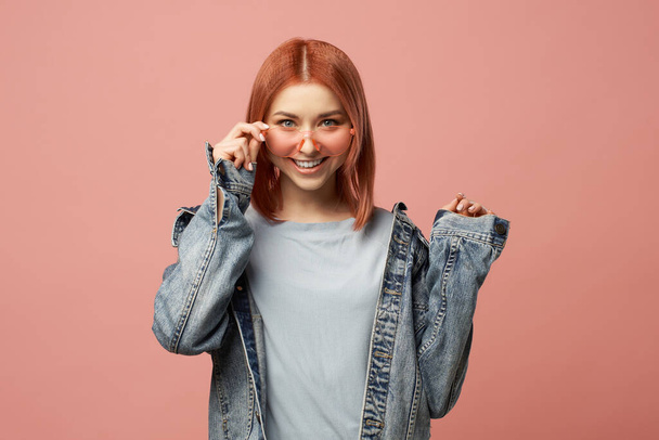 Smiling ginger woman in pink glasses in denim jacket standing in studio - Photo, image
