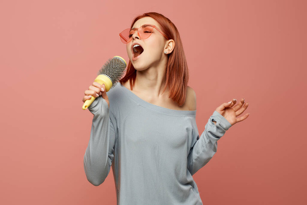 Young singing woman in pink glasses with comb in her hands standing in studio isolated - Foto, Bild