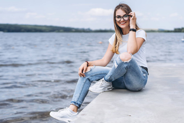 Young woman with long hair in stylish glasses posing on the concrete shore near the lake. Girl dressed in jeans and t-shirt smiling and looking at the camera. - Photo, image