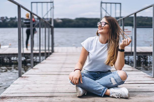 Young woman with long hair in stylish glasses posing on a wooden pier near the lake. Girl dressed in jeans and t-shirt smiling and looking away - Photo, image