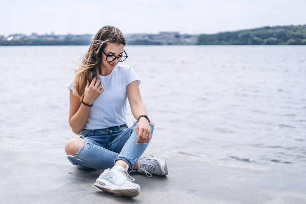 Young woman with long hair in stylish glasses posing on the concrete shore near the lake. Girl dressed in jeans and t-shirt smiling and looking away. - Photo, image