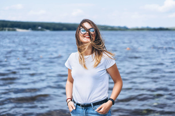 Portrait of a beautiful young woman in stylish glasses. Girl in white tshirt posing on the background of the lake landscape. - Фото, изображение