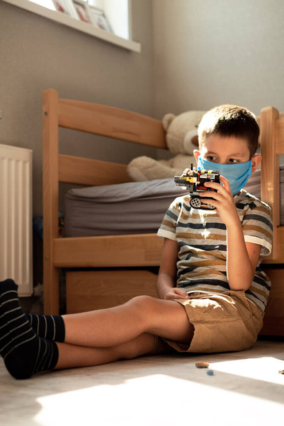 Stay at home quarantine coronavirus pandemic prevention. A boy in a protective medical mask plays and folds the constructor himself in his room. Prevention epidemic. COVID-19. - Фото, изображение