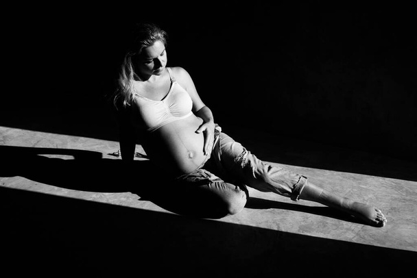 Kaunis raskaana oleva nainen denim haalari ja rintaliivit istuu betonilattialla auringonvalossa. Äitiyden käsite
 - Valokuva, kuva