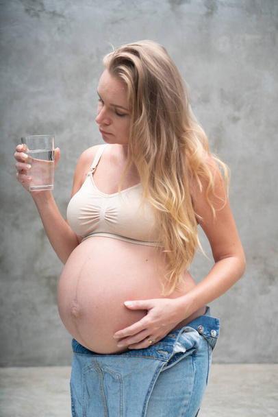Csinos terhes nő farmer kezeslábasban és melltartóban, kezében egy pohár vízzel, miközben a szürke fal fölött áll. Anyaság, terhesség, új élet egészséges étel koncepció - Fotó, kép