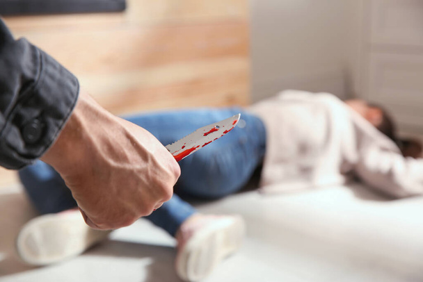 Man with bloody knife and his victim on floor indoors, closeup. Dangerous criminal - Fotoğraf, Görsel