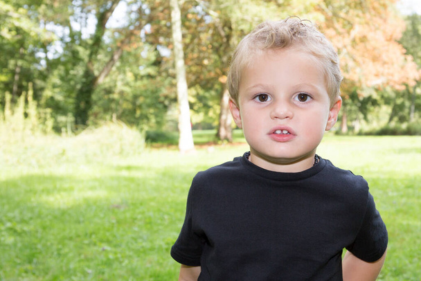 schattig klein blond jongen kind in zwart t-shirt in groen gras veld park - Foto, afbeelding