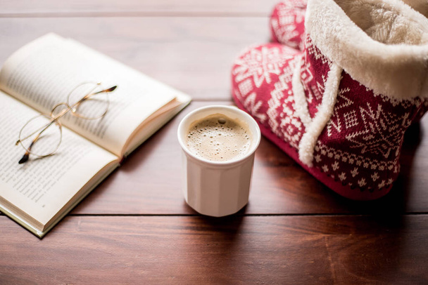 šálek kávy na dřevěném stole se zimními botami a knihou s brýlemi - Fotografie, Obrázek