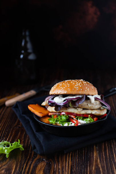 Zdrowy burger dietetyczny z grillowanym filetem z kurczaka, papryką, słodkim ziemniakiem, sałatą, cebulą i jogurtem greckim podawany na patelni na drewnianym stole. Zbliżenie, przestrzeń kopiowania - Zdjęcie, obraz