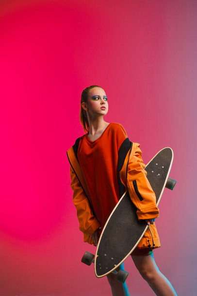 A young girl with bright makeup holds a longboard in her hands on a fashionable neon color background. Portrait of a girl skater - Foto, afbeelding