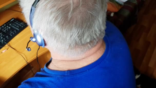 ヘッドフォンを持つ白髪の高齢者は、距離学習のためのコンピュータを使用し、音楽を聞き、 Skypeを介して通信します。現役引退生活、背中からの眺め - 映像、動画