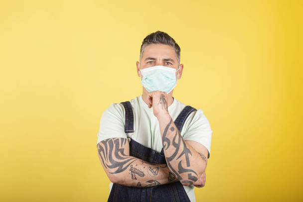 Man with protective medical mask wearing blue denim overalls on yellow background. Health protection, safety and pandemic concept. - Photo, Image