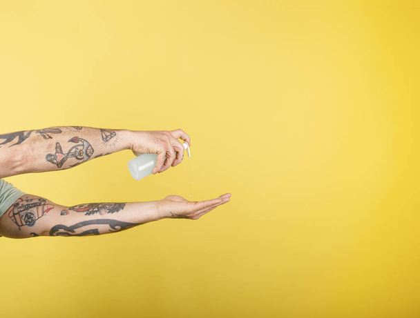 消毒ゲルを使用したスタジオでの入れ墨の男性の腕の肖像画は、黄色の背景にコロナウイルスの予防、単離された画像に対して手を洗う. - 写真・画像