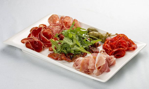 Appetizers of Italian sausages. Prosciutto, chorizo, pancetta and salami. Capers, arugula and sun-dried tomatoes. On white background - Photo, Image