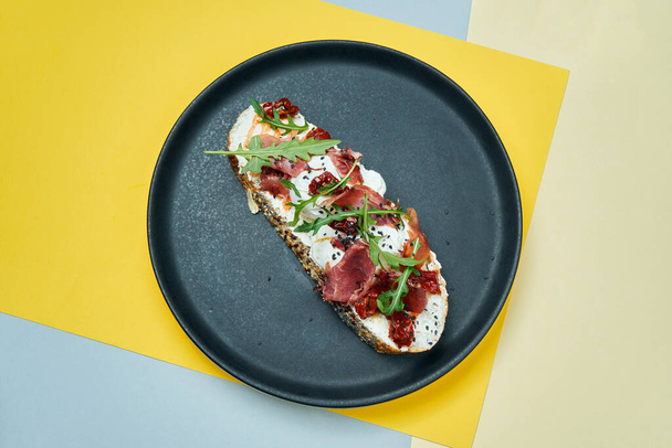 Trendy street snack. Tasty sandwich with jamon, poached eggs, cucumber and microgreen on black plate on colored background. copy space. - 写真・画像