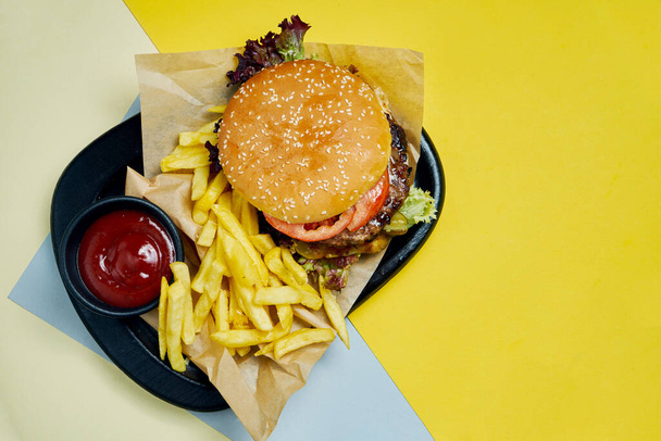 Appetizing American burger with beef, tomatoes, lettuce and fries on a black tray on a colored background. Tasty fast food - Фото, изображение