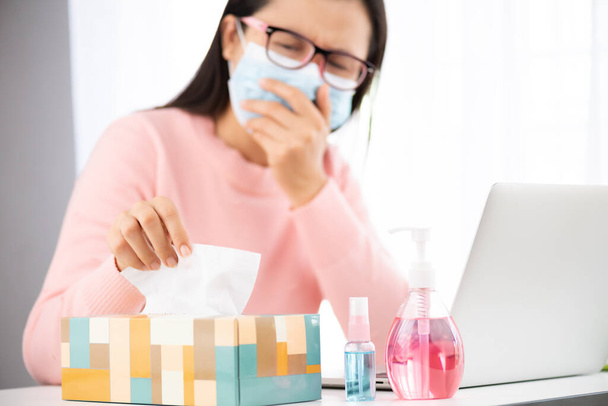 Sick business woman hand taking white tissue paper while cough. Laptop mouse smartphone, sanitizer gel, medical face mask, and alcohol spray for prevention Coronavirus disease (COVID-19). - Фото, зображення