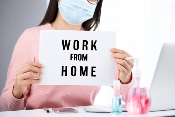 Business woman hand holding white paper with work from home text. Laptop mouse smartphone, sanitizer gel, medical face mask, and alcohol spray for prevention Coronavirus disease (COVID-19). - Fotoğraf, Görsel
