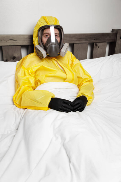 άτομο που αυτο-απομονώνεται στο κρεβάτι φορώντας στολή και μάσκα κινδύνου - Φωτογραφία, εικόνα