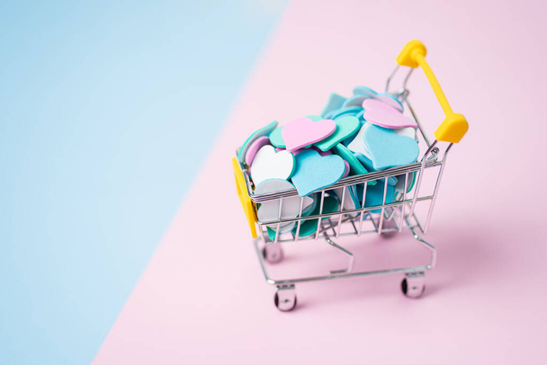 Close-up van een miniatuurkarretje vol kleurrijke hartjes op een tweekleurige roze en blauwe achtergrond met kopieerruimte voor tekst. Valentijnsdag, lente, liefde, kortingen en promoties, online shopping concept - Foto, afbeelding