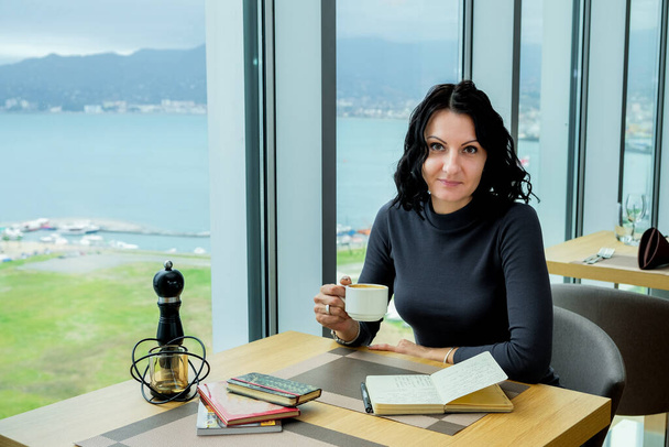 Elegant brunette woman sitting with coffee and notepad. Panoramic window with landscape. Business woman in cozy workplace. Woman taking notes in notepad. - Photo, Image
