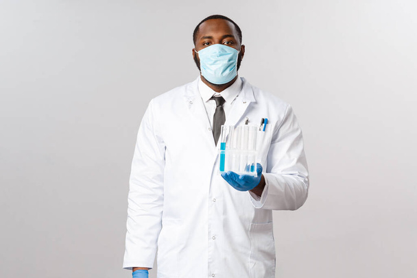 Covid-19, leczenie pacjentów z koronawirusem i koncepcja laboratoryjna. Poważny, profesjonalny, afrykańsko-amerykański lekarz w masce do twarzy i rękawicach lateksowych, trzymający fiolki, probówki ze szczepionką - Zdjęcie, obraz