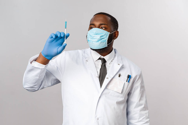 Pokryty19, pandemia i koncepcja opieki zdrowotnej. Poważny, przystojny, afrykańsko-amerykański lekarz w masce na twarz, masce lateksowej, patrząc na strzykawkę ze szczepionką przeciw wirusowi, lecząc pacjentów, szare tło - Zdjęcie, obraz
