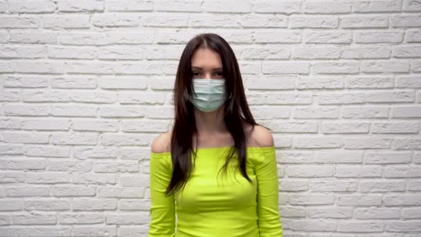 Cute young brunette woman in green jacket in protective medical mask on face feels a sense of fear against white brick wall background. She is scared by the corona virus. Horror captures the heart. - Footage, Video