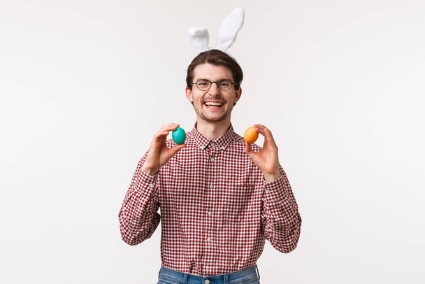Tradycje, święta religijne, koncepcja świętowania. Zabawny i uroczy brodaty mężczyzna spędza czas z rodziną podczas Wielkanocy, nosi królicze uszy, trzyma dwa malowane jajka i uśmiecha się radośnie - Zdjęcie, obraz