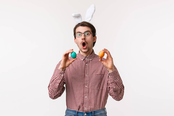 Traditions, religious holidays, celebration concept. Funny joyful young man with beard in glasses, wear cute rabbit ears and hold two painted eggs, playing game on Easter day, white background - Fotografie, Obrázek