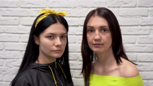 Zwei hübsche kaukasische Mädchen mit langen dunklen Haaren schauen ernst in die Kamera. Schöne Frau in grünem Sakko lacht, lächelt, redet und grimmt vor weißem Backsteinhintergrund.  - Filmmaterial, Video