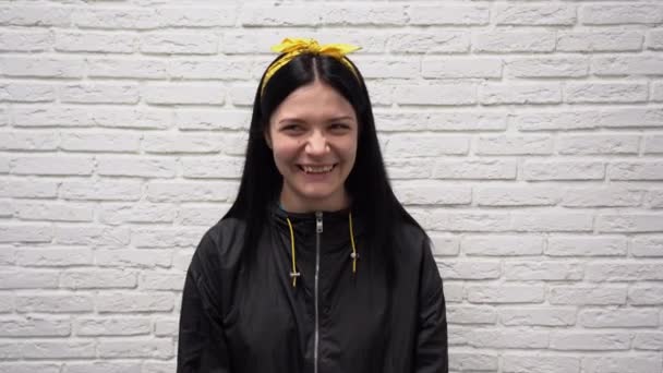 Portrait de femme caucasienne mignonne heureuse en veste noire avec les cheveux longs et bandeau jaune. Belle jeune fille rit et sourit sur fond de mur de brique blanche gros plan
. - Séquence, vidéo