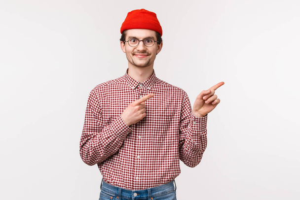 Талия портрет удовлетворенный счастливый улыбающийся молодой хипстер парень в красной шапочке, очки указывая пальцами правую верхнюю рекламу с приятным выражением лица, нашел отличный продукт или обслуживание компании
 - Фото, изображение