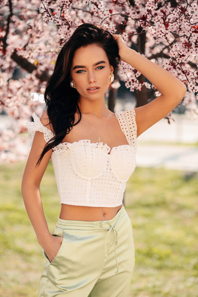 fashion outdoor photo of beautiful girl with dark hair in elegant dress posing among blossoming peach trees in garden - Fotoğraf, Görsel
