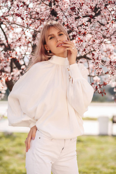 fashion outdoor photo of beautiful girl with blond hair in elegant dress posing among blossoming peach trees in garden - Photo, Image