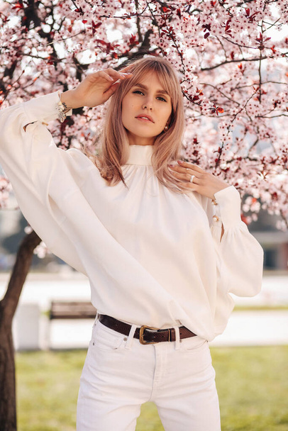 fashion outdoor photo of beautiful girl with blond hair in elegant dress posing among blossoming peach trees in garden - 写真・画像