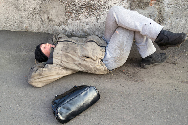 Бездомна брудна жінка лежить під стіною і спить на землі. Жіноча сумка неподалік
 - Фото, зображення