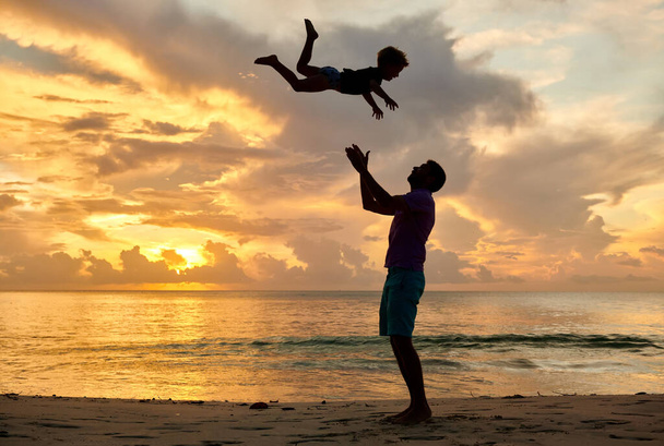 Iloinen lapsi, isä oksentaa pojan ilmaan. Kolmivuotias pikkupoika rannalla isän kanssa auringonlaskun aikaan. Kesäloma Seychelleillä, Mahe
. - Valokuva, kuva