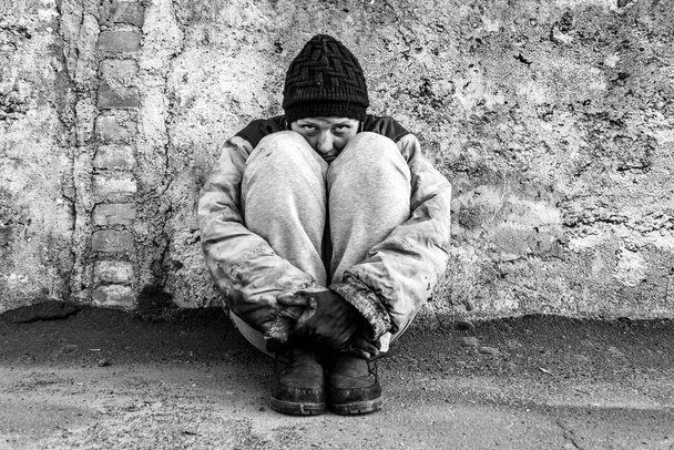 Mujer sucia sin hogar sentada bajo una pared gris abrazando sus piernas
 - Foto, imagen