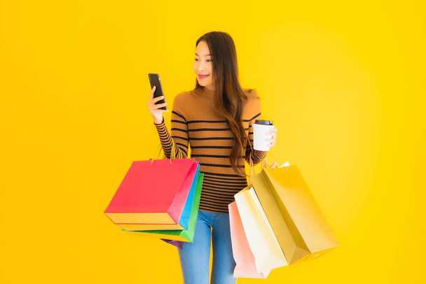 Hermosa mujer asiática joven utilizar teléfono móvil inteligente o teléfono celular con taza de café y bolsa de compras de color sobre fondo aislado amarillo
 - Foto, Imagen