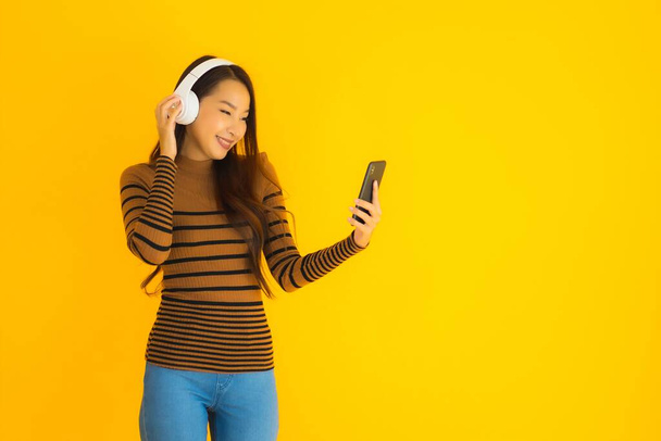 Muotokuva kaunis nuori aasialainen nainen käyttää älypuhelimella tai matkapuhelimella kuulokkeella kuunnella musiikkia ja tanssia keltaisella eristetyllä taustalla
 - Valokuva, kuva