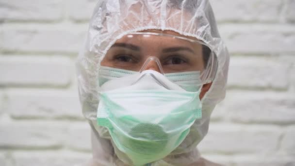 Caucasian man wear protective suit, respirator, glasses and medical mask from virus and hood on head. Cute girl winks right eye twice on a white brick wall background close up. - Metraje, vídeo