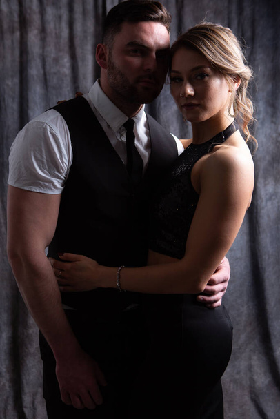 Sexy beautiful stylish couple: a charming blonde girl in a black dress and a brutal man in a suit stand gently hugging in a dark Studio under the weak light of spotlights - Photo, Image