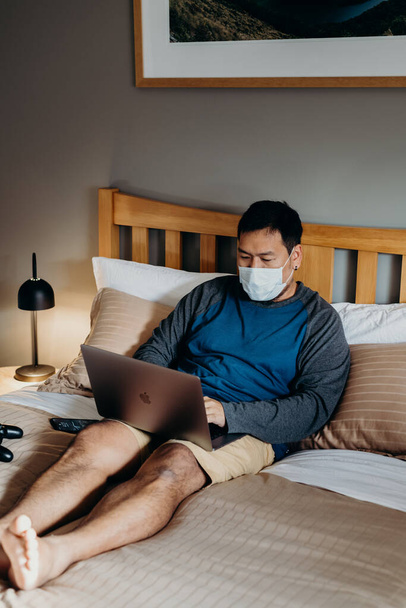 Hombre asiático con máscara médica. Sentarse y usar el portátil en el dormitorio
. - Foto, imagen