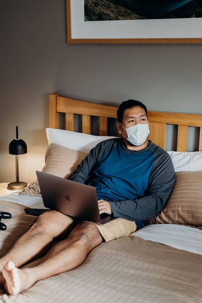 Hombre asiático con máscara médica. Sentarse y usar el portátil en el dormitorio
. - Foto, Imagen