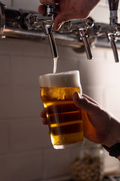 Egy férfi könnyű arany sört tölt egy pohárba egy sörcsapból, fehér sörhabbal öntve, közelről. - Fotó, kép