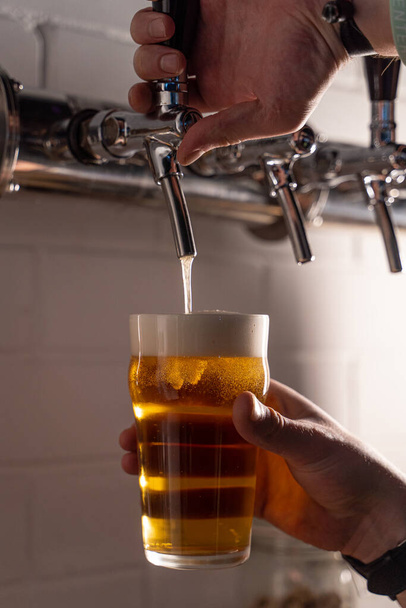 Άνθρωπος ρίχνει ελαφριά χρυσή μπύρα σε ένα ποτήρι από μια βρύση μπύρα, ρίχνουμε με λευκό αφρό μπύρα, κλείστε τα χέρια - Φωτογραφία, εικόνα