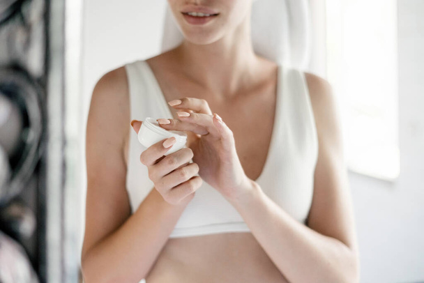 Gewassen uitzicht van jonge volwassen vrouw met biologische gezichtscrème cosmetische in de handen, in de buurt van spiegel in witte badkamer, het aanbrengen van lotion op de huid - Foto, afbeelding