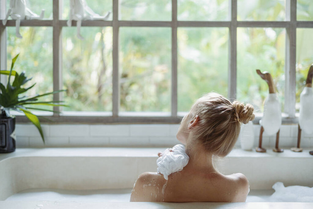 Skin care concept. Back view of young adult woman taking bath, washed shoulder, holding sponge in hand and lying in foam water, spending morning in bathroom - Photo, Image